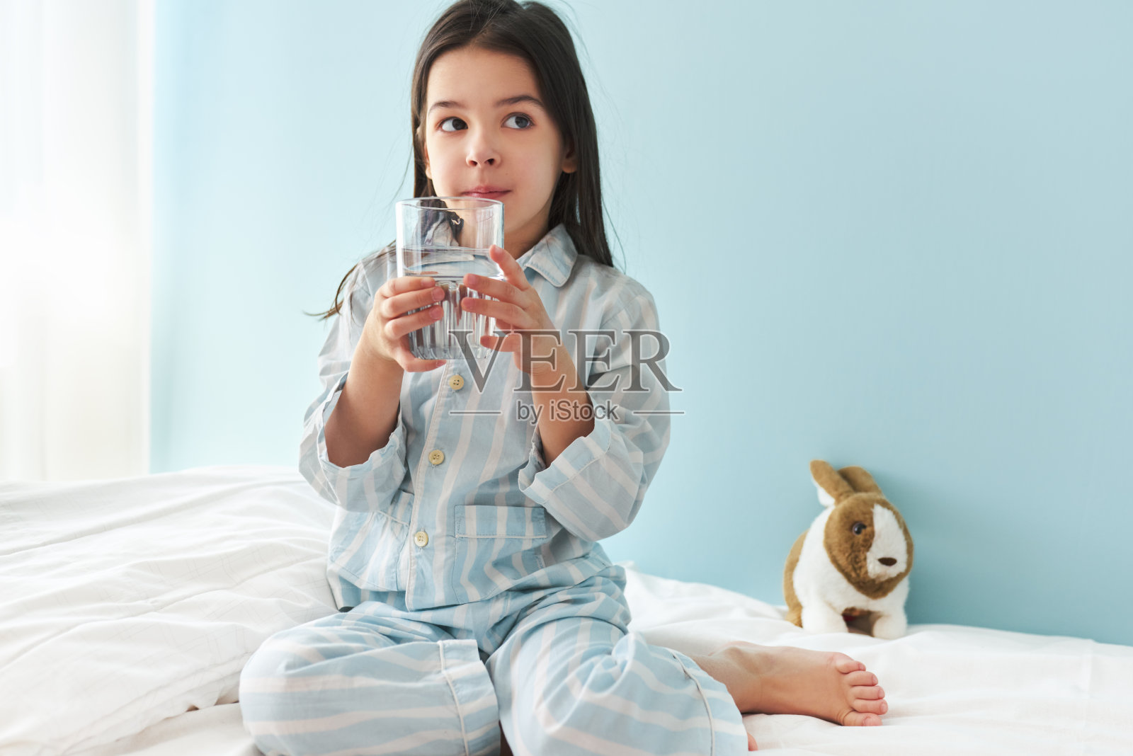 漂亮的小女孩在家里的床上喝着淡水。可爱的孩子在早上拿着一杯纯净水。健康的生活方式的概念。照片摄影图片