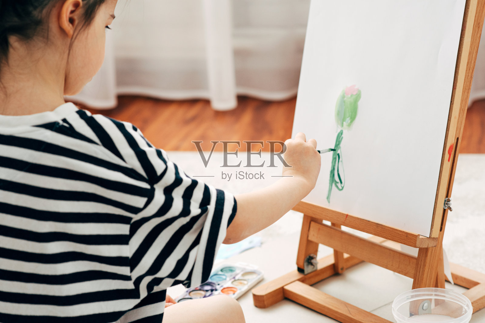 一个小女孩在家里画架上画花的后视图。可爱的孩子坐在地毯上，在她的房间里画画。照片摄影图片