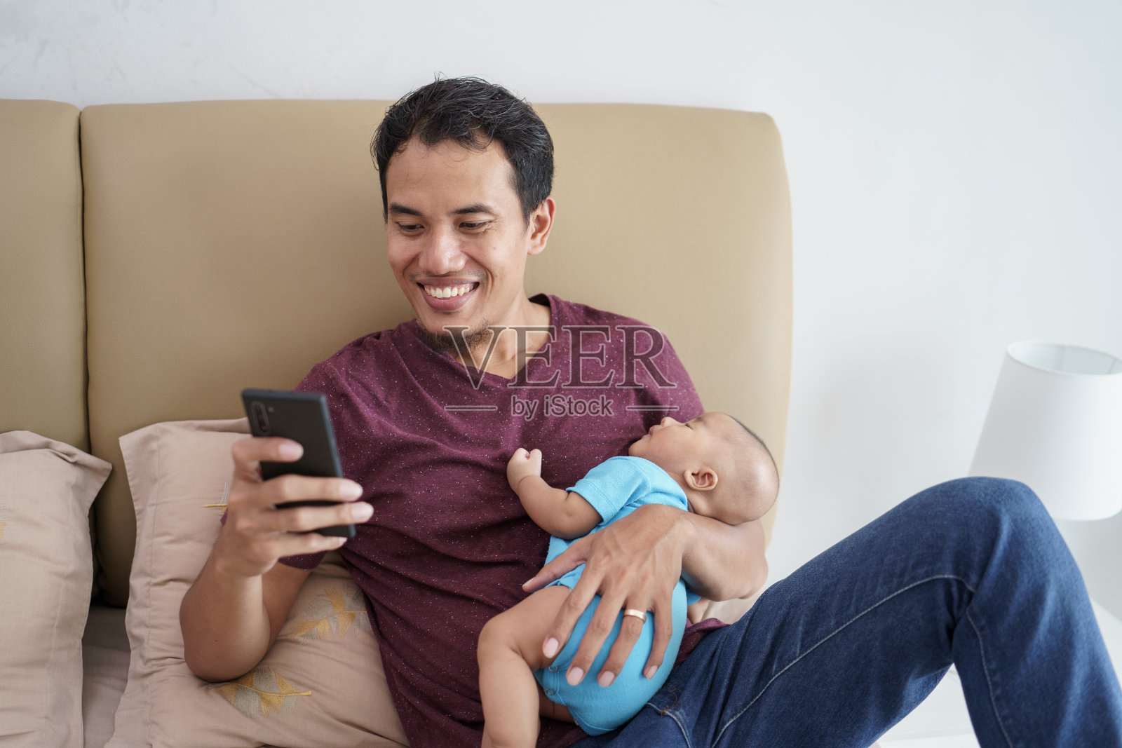 父亲抱着他刚出生的可爱宝宝在他的胳膊上睡觉，同时在床上玩手机照片摄影图片