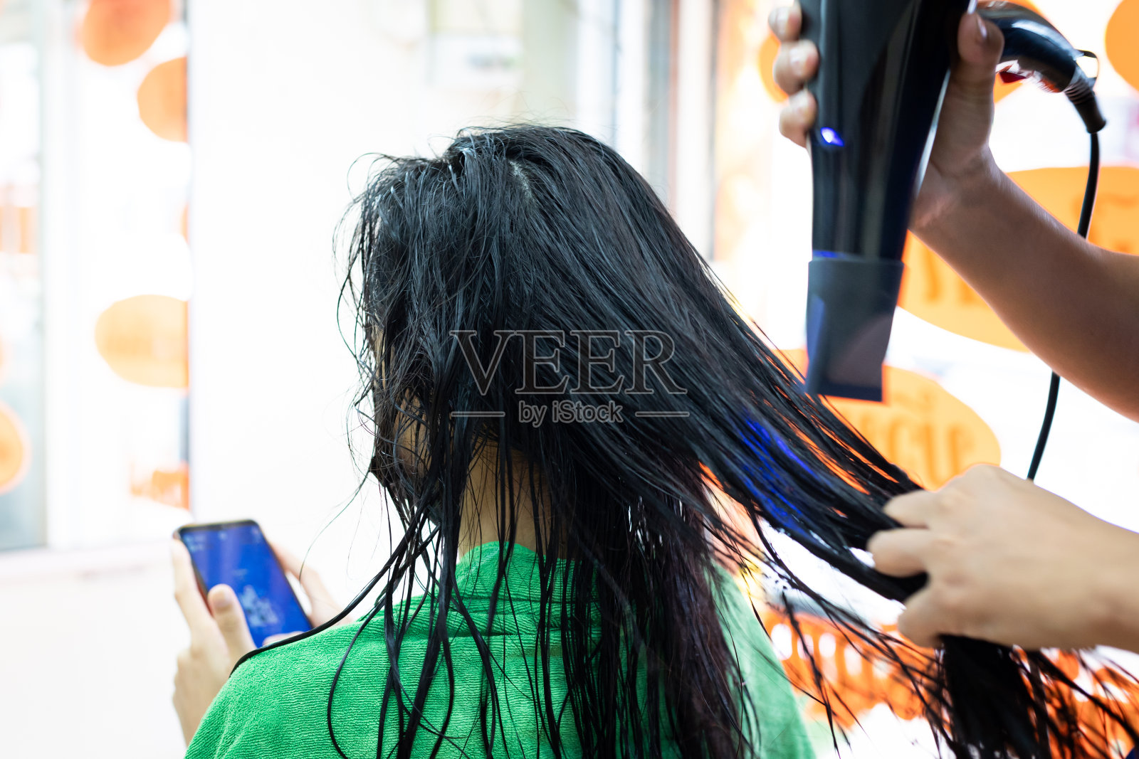 一名年轻的亚洲女子戴着防护面具，在美发师用吹风机吹头发时使用智能手机。美发师在发廊为顾客烘干头发。照片摄影图片