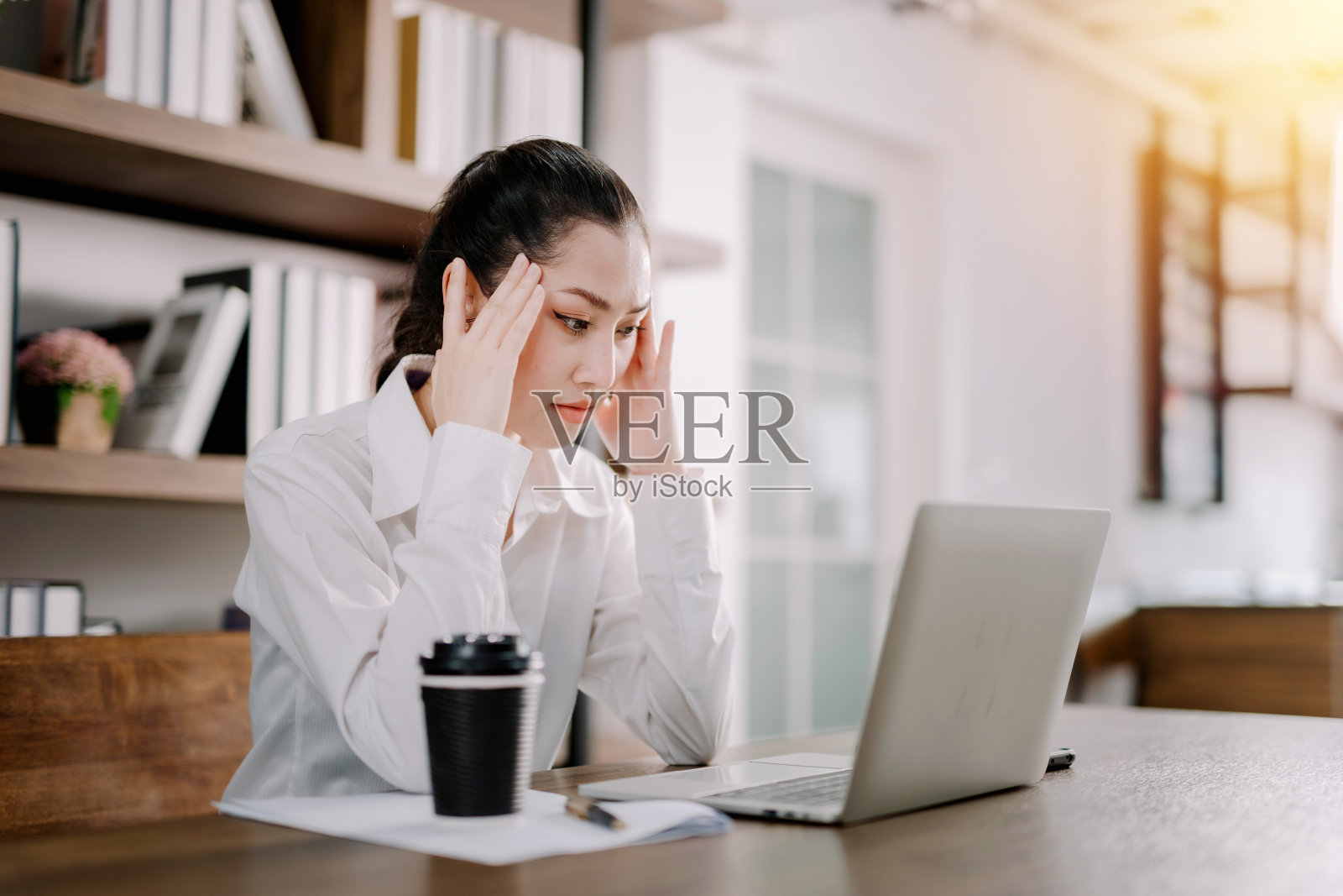 一位漂亮的女商人坐在她的办公桌前，手里拿着一台笔记本电脑，她的压力很大，头痛的手踢着她的头，工作中的压力和无聊的概念。照片摄影图片