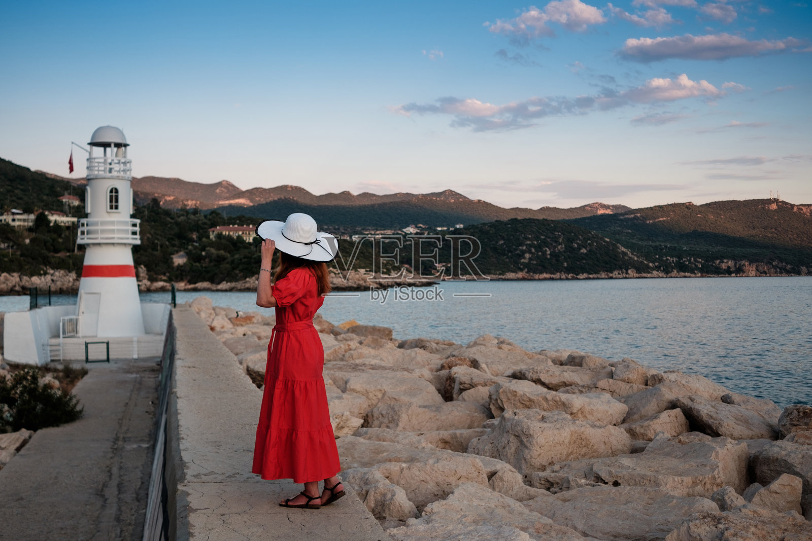 这是一幅美丽的画，画中的女孩戴着帽子，穿着红色的衣服，在日落时靠近灯塔和船只。土耳其安塔利亚卡斯老城港。照片摄影图片