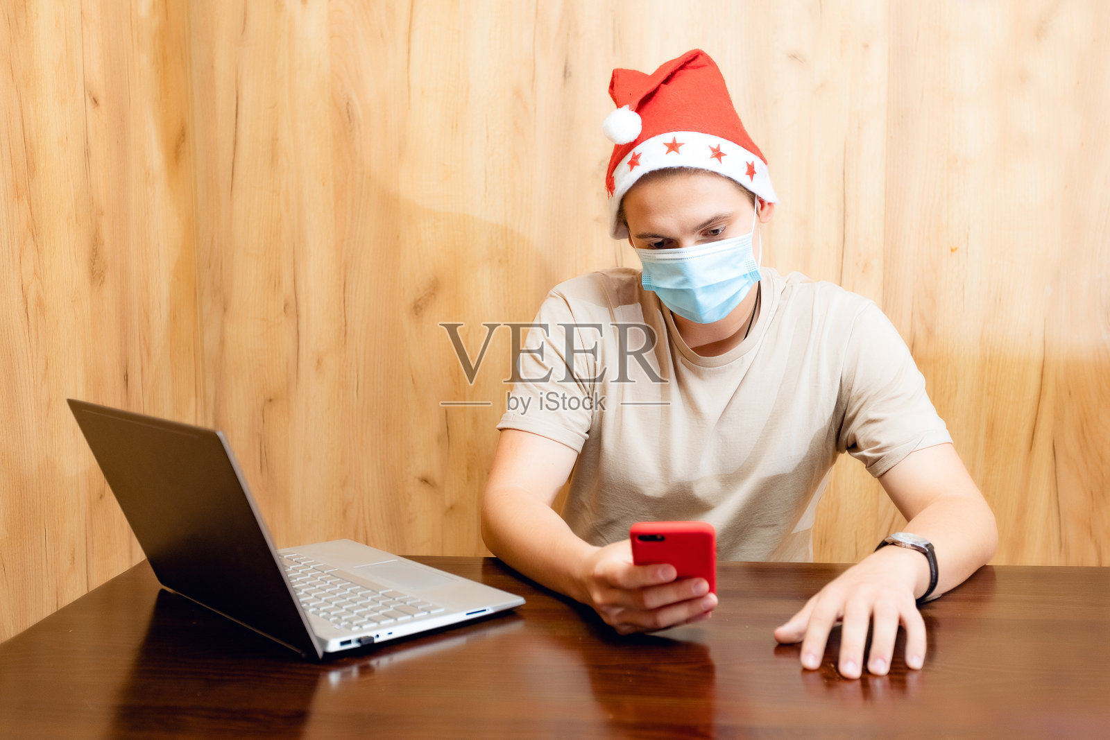圣诞男性肖像，戴着红色圣诞帽和面部保护口罩的男子在大流行冠状病毒期间使用笔记本电脑进行视频通话，新年概念。有人在用手机打电话照片摄影图片