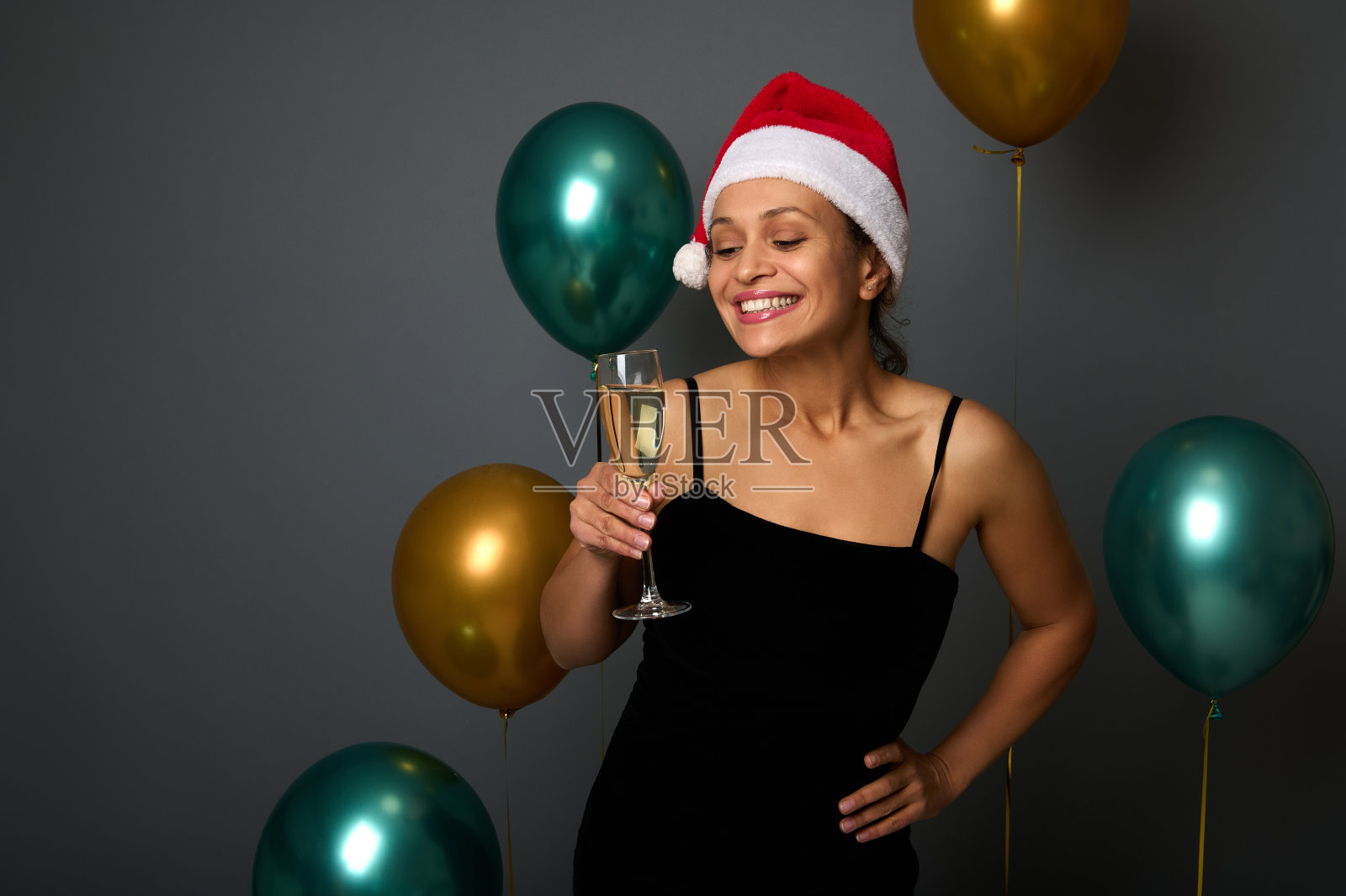 有魅力的女人戴着圣诞帽，身穿黑色礼服，在圣诞派对上玩得很开心，微笑着拿着香槟长笛看着镜头，在灰色背景下与闪闪发光的金绿色气球隔离照片摄影图片