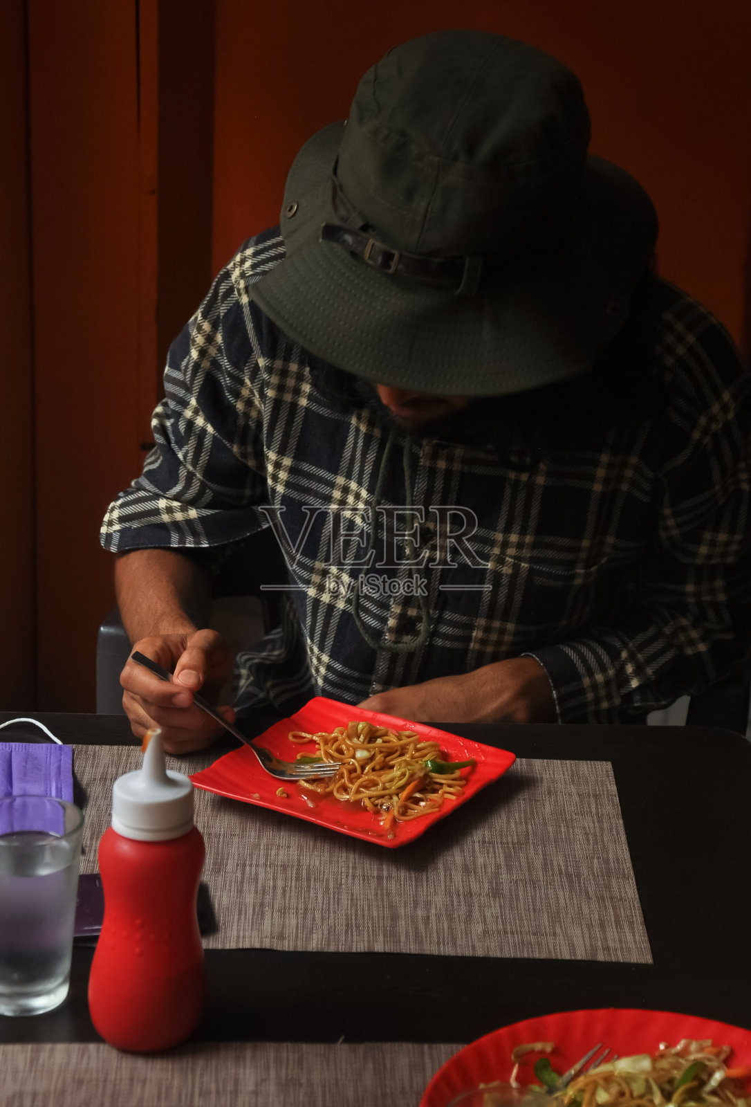 新冠肺炎大流行期间，一名留胡子和长发的年轻人在餐厅吃面条。一个男人戴着乡下帽吃快餐照片摄影图片