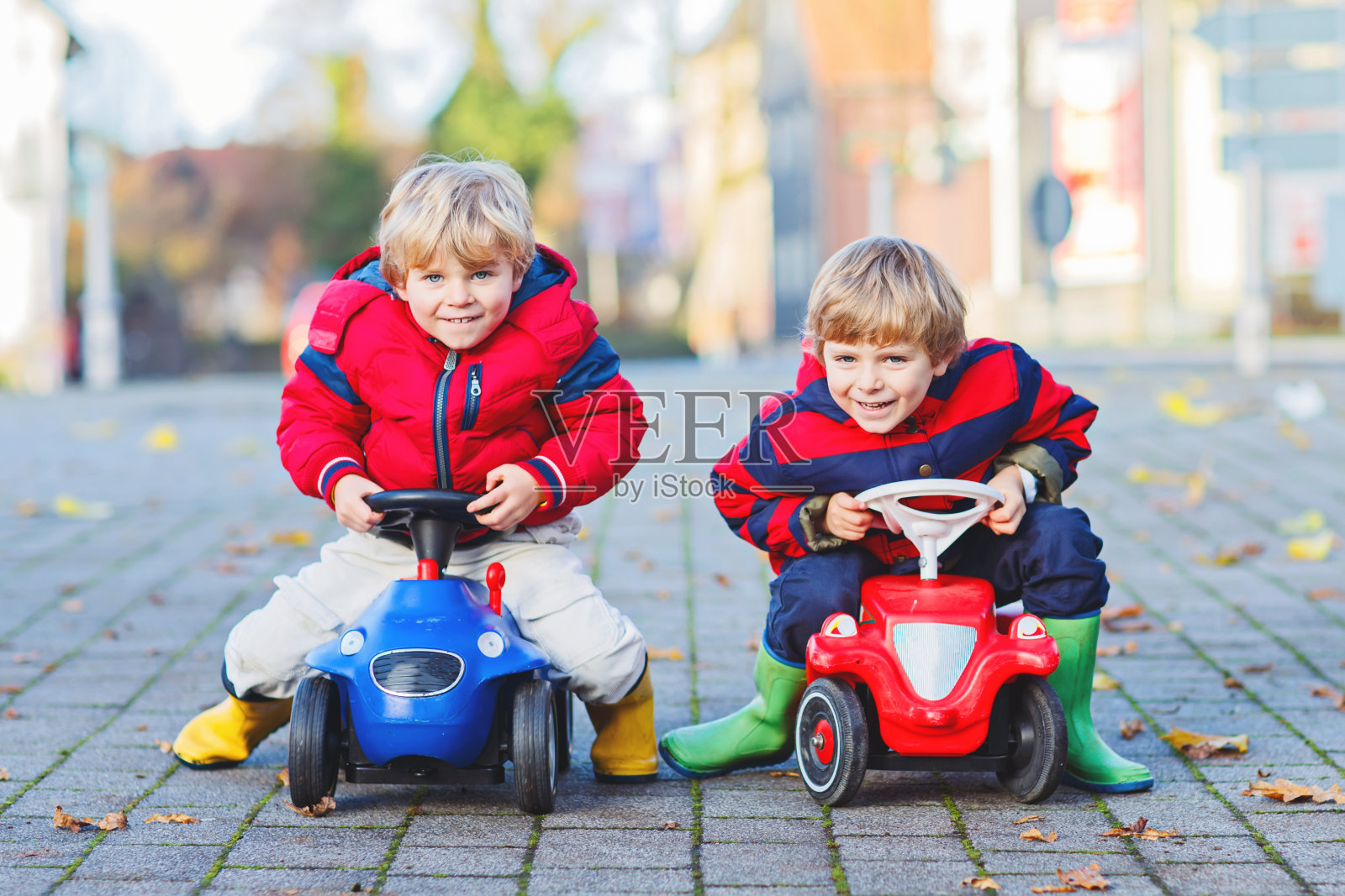 两个小男孩穿着五颜六色的衣服和雨靴开着玩具车。双胞胎在户外竞争。秋日儿童积极休闲活动。照片摄影图片