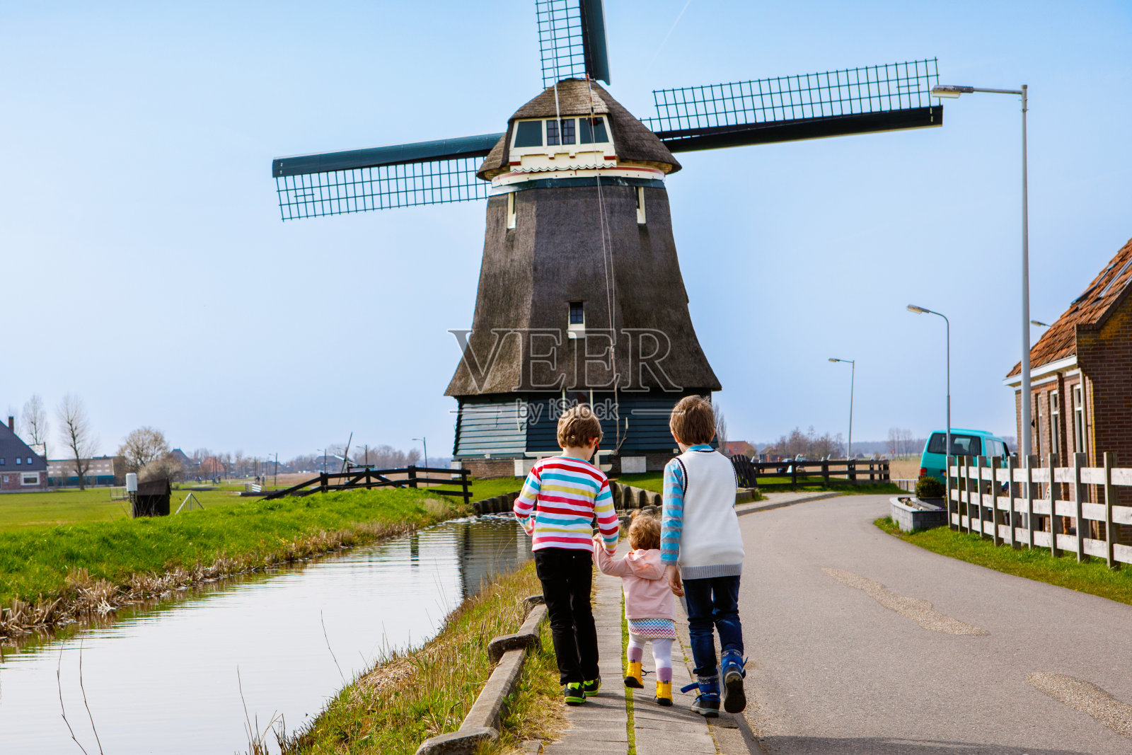 在荷兰，两个男孩和一个小女孩走在有大风车的街道上。一家三个孩子在荷兰度假。幸福的兄弟姐妹手牵着手。照片摄影图片