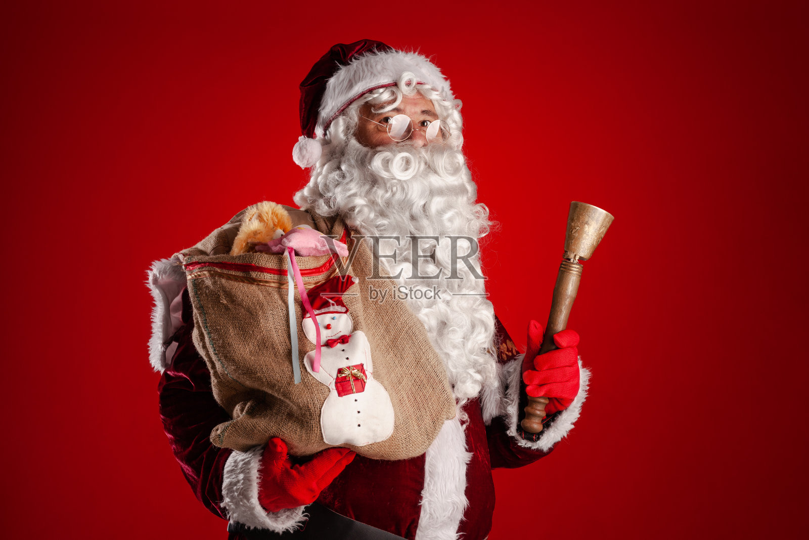 圣诞老人拿着袋子和铃铛的肖像衬着红色的背景。照片摄影图片