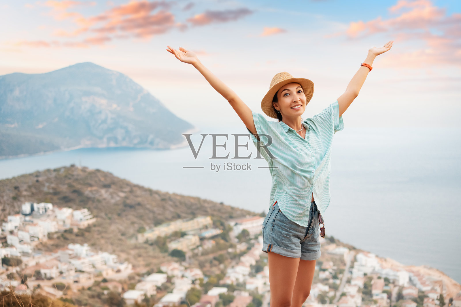 快乐的度假者戴着帽子，张开双臂站在观景台上，欣赏着地中海海岸度假小镇的全景照片摄影图片