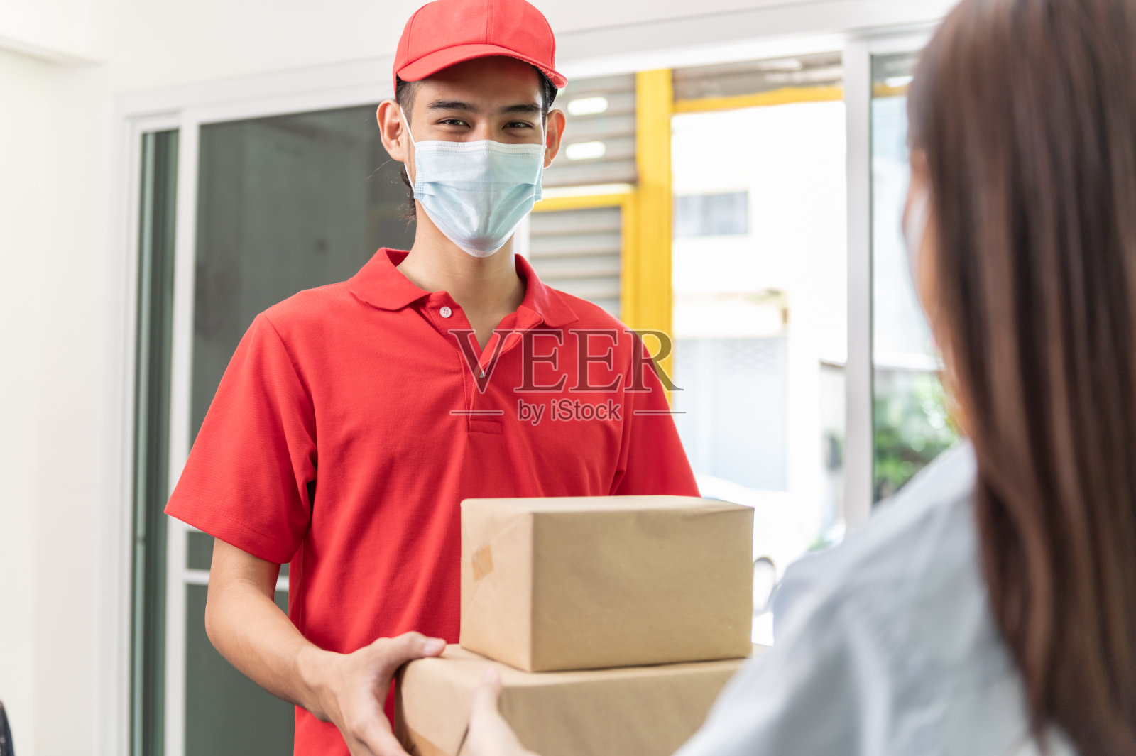 年轻的亚洲女性戴着口罩，在家里接收快递公司员工的包裹和邮件，以预防冠状病毒感染。照片摄影图片